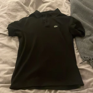 Svart pikétröja från Lacoste - Klassisk svart pikétröja från Lacoste med korta ärmar och ikoniska krokodilloggan på bröstet. Tröjan har krage och knäppning framtill. Perfekt för en clean och sportig stil, passar lika bra till jeans som till shorts.