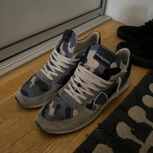 Philippe Model sneakers med camomönster - Snygga sneakers från Philippe Model med grå mockadetaljer och blått, svart och beige camouflagemönster. Skorna har vita snören, rund tå och platt sula. Perfekta för dig som vill sticka ut med en cool streetstyle.