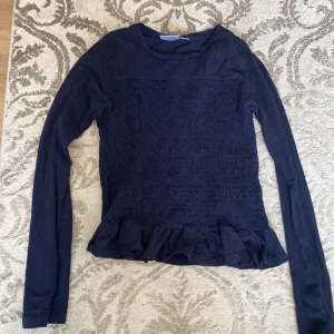 Gina tricot Mörkblå långärmad tröja - Supersöt mörkblå långärmad topp från Gina Tricot. Smockad överdel och volangkant nedtill ger en trendig vibe. Mjuk bomullskänsla och perfekt passform för dig som gillar stilrena men ändå lekfulla detaljer.