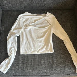 Vit långärmad topp med cut out från Divided - Säljer en vit långärmad topp från Divided med snygg cut out-detalj över bröstet och twistad axel. Toppen är i mjuk bomull och har en figurnära passform som ger en modern vibe. Perfekt att styla med jeans eller kjol för en trendig look.