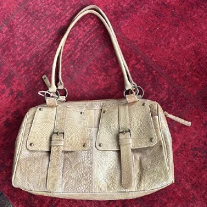 Beige axelväska i skinn med ormmönster - Snygg beige axelväska i skinn med ormmönstrad yta och två stora fickor framtill med spännen. Väskan har dubbla handtag och dragkedja upptill. Perfekt för dig som vill ha en unik och rymlig väska med coola detaljer.