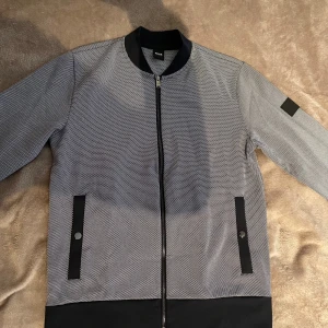 Hugo Boss Bomberjacka - En snygg Boss bomberjacka med en dragkedja framtill och två fickor på sidorna. Den har en klassisk bomberjacka design med en ribbstickad krage, manschetter och nederkant. Ny pris 310 euro! Använd bara 1 gång!
