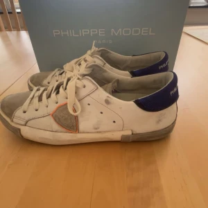 Philippe Model vita sneakers med blå detalj - Snygga vita sneakers från Philippe Model med grå mockadetaljer på tån och sidan, samt blå mesh vid hälen. Skorna har snörning och en orange kant runt loggan på sidan. Perfekta för dig som gillar en stilren men ändå unik look.               Storlek 43 och priset går att diskutera