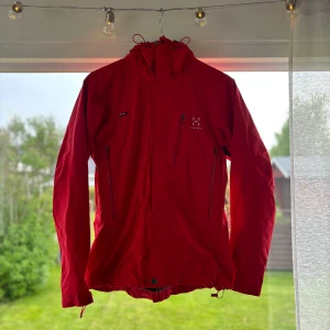 Röd vindjacka från Haglöfs dam S - Riktigt snygg röd vindjacka från Haglöfs i dammodell, storlek S. Jackan har huva, flera dragkedjeförsedda fickor och justerbar nederkant. Perfekt för uteliv och äventyr, med stilren design och logga på bröstet.