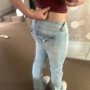 True Religion Ljusblå bootcut jeans  - Säljer ett par ljusblå jeans från True Religion med bootcut passform och låg midja. Jeansen har slitna detaljer framtill. Ganska använda men är i väldigt bra skick. Dock är en knapp borta på höger fickan!