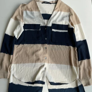 Randig skjorta från Zara  - Snygg långärmad skjorta från Zara Basic med breda, målade ränder i beige, marinblått och vitt. Skjortan har v-ringning, stora bröstfickor och knappar framtill. Materialet är lätt och har en skön struktur. Perfekt för dig som gillar en avslappnad men trendig look.