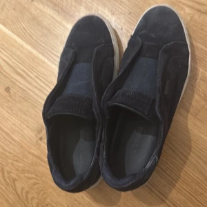 Svarta sneakers från Axel Arigato - Snygga svarta slip-on sneakers från Axel Arigato med vit sula och diskret logga i guld på sidan. Ovandelen är i mocka och textil, med bred resår över vristen för enkel påtagning. Perfekta för en clean och stilren look.