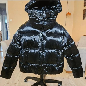 Svart pufferjacka från Calvin Klein Jeans - Snygg svart pufferjacka från Calvin Klein Jeans med glansig finish och stor huva. Jackan har dragkedja och knappar framtill samt stora fickor. Perfekt för kalla dagar när du vill hålla stilen och värmen. Den är stor i storleken.