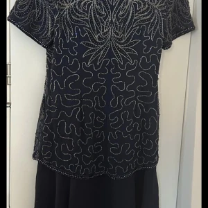 Mörkblå pärlbroderad klänning Carolyne Barton - Supersnygg mörkblå klänning med v-ringning och korta ärmar från Carolyne Barton. Klänningen är täckt av glittriga pärlbroderier i silver och har dragkedja bak. Perfekt för dig som vill sticka ut med eleganta detaljer och unik design.