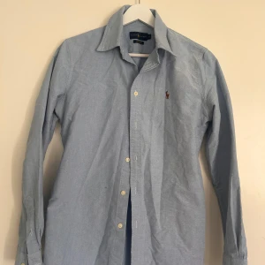 Ljusblå skjorta från Ralph Lauren S - Klassisk ljusblå skjorta från Ralph Lauren i custom fit. Skjortan har lång ärm, knappar framtill och den ikoniska broderade polospelaren på bröstet. Perfekt för en clean och stilren look. Tillverkad i mjuk bomull för skön känsla hela dagen.