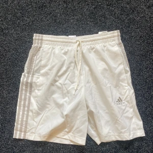 Vita Adidas shorts med snörning M - Snygga vita Adidas shorts med klassiska tre ränder längs sidorna och logga framtill. De har elastisk midja med snörning och är tillverkade i lätt polyester med meshfoder. Perfekta för träning eller chill dagar på sommaren.