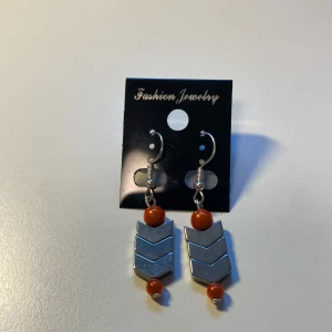 Unika Silvriga örhängen med orange pärlor *Endast 1 Kvar* - Unika örhängen med silvriga, pilformade detaljer och glansiga orange pärlor. Snyggt geometriskt uttryck som sticker ut och ger en cool vibe till din stil. Perfekta för dig som gillar att mixa färg och metall i dina accessoarer.