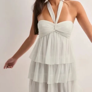 Vit halterneckklänning med volanger - Supersöt vit klänning med halterneck och flera lager volanger. Omlottkänsla upptill med twistad detalj vid bysten. Lätt och luftig, perfekt för varma dagar eller sommarens alla tillfällen.