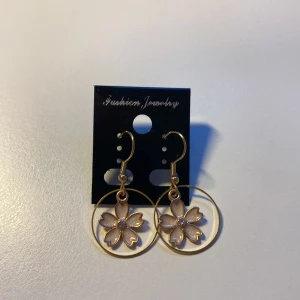 Handgjorda Guldiga örhängen med blomma - Snygga örhängen i guldton med hängande blommor inuti tunna cirklar. Blommorna har fem kronblad i ljusrosa och en liten glittrig sten i mitten. Perfekta för att ge din look en söt och trendig touch.