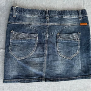 Mörkblå jeanskjol med nitar - Snygg mörkblå jeanskjol i kort modell med ”stenar” på fickorna fram och bak. Klassisk femficksdesign, knapp och dragkedja framtill samt bälteshällor. Midjemått: 34,5 cm rakt över. Längd: 31 cm. Stretchig. Skriv gärna om ni har några frågor. OBS dubblett av tidigare annons för fri frakt