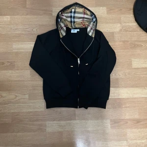 Svart hoodiejacka från Burberry - Svart hoodiejacka från Burberry med dragkedja och klassisk rutig huva i beige, rött och svart. Jackan har snörning i huvan och är tillverkad i mjuk bomull. Perfekt för dig som vill ha en stilren och lyxig streetlook.