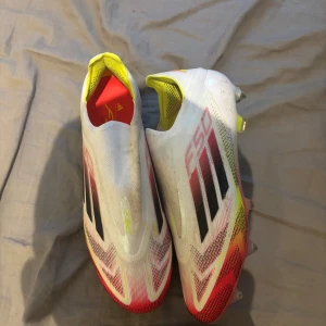 Adidas F50 vita fotbollsskor - Snabba Adidas F50 fotbollsskor i vitt med svarta och röda detaljer samt gula inslag. Skorna har en strömlinjeformad design utan snörning, metall-dobbar och mesh-liknande ovandel för lätt känsla på planen. Perfekta för dig som vill sticka ut på matchen.