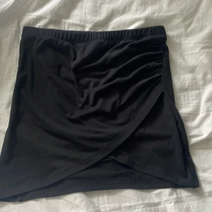 Svart omlottkjol från BikBok - Svart kortkjol från BikBok i mjukt bomullsmaterial. Kjolen har omlottdesign framtill och resår i midjan för en bekväm passform. Lite nopprar på en av sidorna och inuti vid midjan men inget som syns tydligt. Säljer då den är förstor