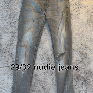 Grå slitna jeans från Nudie Jeans - Säljer ett par grå slitna jeans från Nudie Jeans med coola slitningar och hål på ena benet. Jeansen har en smal passform och klassisk femficksdesign. Perfekta för dig som gillar en edgy streetstil.