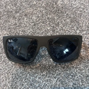 Svarta Ray-Ban solglasögon med blå detalj - Snygga svarta Ray-Ban solglasögon med rektangulära bågar och coola blå detaljer på skalmarna. Mattsvart finish och mörka glas ger en modern vibe. Perfekta för dig som vill sticka ut med en sportig och stilren look.