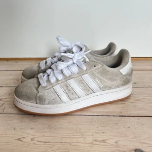 Adidas Campus sneakers i beige mocka - Snygga Adidas Campus sneakers i beige mocka med klassiska vita ränder och tjocka vita snören. Skorna har en låg profil, vit sula och diskret Adidas Campus-tryck på sidan.