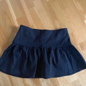 Svart randig kortkjol med dragkedja - Supersöt svart kortkjol med tunna vita ränder och volang nedtill. Kjolen har en markerad midja och en diskret dragkedja i sidan för enkel på- och avtagning. 