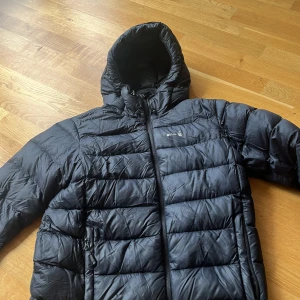 Svart pufferjacka från Sail racing - Svart pufferjacka från Sail racing med huva och dragkedja framtill. Jackan har quiltad design och två fickor med dragkedja. Perfekt för kalla dagar när du vill hålla värmen och ändå se stilren ut. Den är lite sliten på vissa ställen men inga hål 