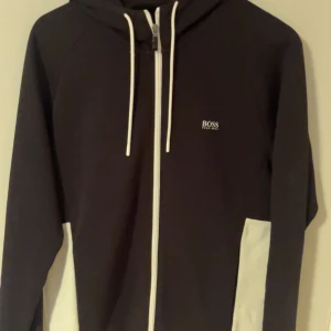 Svartvit hoodie från BOSS - Snygg svart hoodie från BOSS med vita detaljer och logga på både bröstet och ryggen. Tröjan har hel dragkedja, huva med vita snören och coola kontrastpaneler i vitt längs sidorna och på ryggen. Perfekt för en stilren och sportig look.