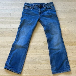 Blå raka jeans från Diesel - Snygga blå jeans från Diesel med klassisk femficksdesign och raka ben. Jeansen har tydliga slitningar och detaljer som Diesel Industry-märke på fickan och läderpatch bak i midjan. Perfekta för en avslappnad look.