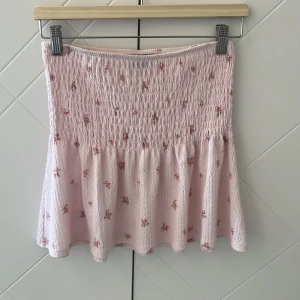 Ljusrosa blommig kortkjol med volang - Supersöt ljusrosa kortkjol med små blommor i rosa och grönt. Passar också att använda som en topp!!💕 Säljer så jag inte fått någon användning av den!