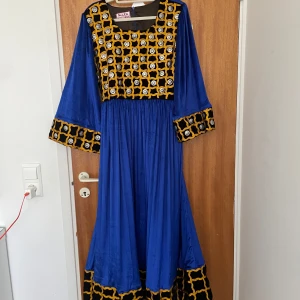 Blå långklänning med broderade detaljer - Unik blå afghansk klänning från Nilla Boutique med vida ärmar och broderade detaljer i svart, gult och silver på bröst, ärmslut och nederkant. Klänningen har en lös passform och är tillverkad i ett glansigt material som ger en lyxig känsla.