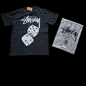 Svart Stüssy t-shirt med tärningsprint - Svart t-shirt från Stüssy med ikonisk logga på bröstet och stort tryck av två vita tärningar på ryggen. Klassisk passform och rund halsringning. Tillverkad i 100% bomull för skön känsla. Storlek S. Mitt pris 250 kr. Det här är en gaaaaalen deal för dig som vill ha en riktigt fet t-shirt.