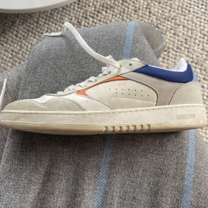 Axel Arigato sneakers i vitt och blått - Snygga sneakers från Axel Arigato i vitt skinn och mocka med blå och orange detaljer. Klassisk låg modell med rund tå, snörning och platt sula. Perfekta för dig som gillar stilrena men ändå unika sneakers.