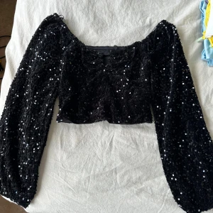 Svart paljettopp från new yorker - Glittrig svart topp med långa puffärmar och croppad passform. Hela toppen är täckt av svarta paljetter som reflekterar ljuset snyggt. Perfekt för dig som vill sticka ut med en statement-piece. Säljes i storlek M.