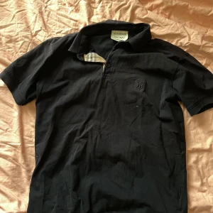 Svart Burberry pikétröja  - Snygg svart t-shirt från Burberry med klassisk krage och korta ärmar. Har broderad Burberry-logga på bröstet och ikoniskt rutigt tyg vid knäppningen. Tillverkad i mjuk bomull, perfekt för en stilren och clean look.