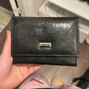 Svart plånbok i skinn från Ripani - Snygg svart plånbok från Ripani i äkta skinn med glansig finish och präglat ormmönster. Plånboken har flera fack för kort och mynt, samt en stilren metallogo framtill. Perfekt storlek för att ha med sig överallt.