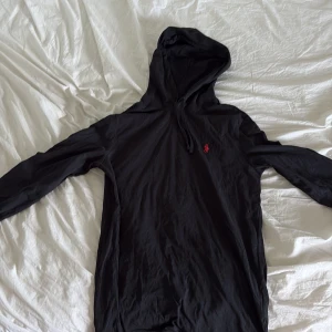 Svart långärmad tröja från Polo Ralph Lauren - Svart långärmad tröja från Polo Ralph Lauren med klassisk röd broderad logga på bröstet. Tröjan har huva med snörning. Långärmad och perfekt för en chill och stilren look.