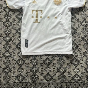 Bayern München Mané tröja Adidas 152 - Vit Bayern München fotbollströja från Adidas med guldiga detaljer och tryck. Kortärmad modell med klubbmärke, sponsorlogga och Mané 17 på ryggen. Tillverkad i lätt och ventilerande material, perfekt för match eller träning.