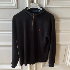  Ny Svart half zip tröja Polo Ralph Lauren - Svart långärmad tröja från Polo Ralph Lauren med half zip och röd broderad logga på bröstet. Tillverkad i mjuk bomull, perfekt för lager-på-lager. Klassisk och stilren design med normal passform och hög krage.