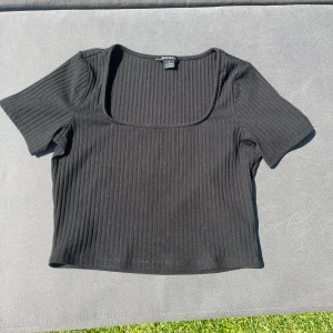 Svart ribbad topp från Monki - Svart croppad topp från Monki med fyrkantig ringning och korta ärmar. Toppen är ribbad och har en tight passform som ger en clean och stilren look. Perfekt att matcha med jeans eller kjol för en trendig vibe.