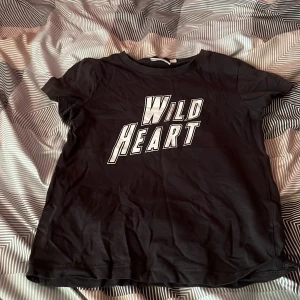 Svart t-shirt Wild Heart KappAhl - Svart t-shirt från KappAhl med coolt tryck 'Wild Heart' i vitt framtill. Klassisk rund halsringning och korta ärmar. Skön passform och mjukt bomullsmaterial, perfekt till jeans eller shorts för en avslappnad vibe.