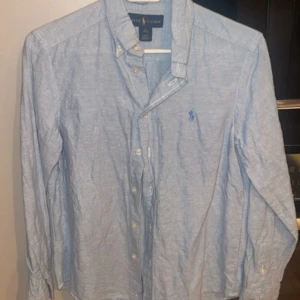 Ljusblå skjorta från Ralph Lauren - Säljer en klassisk ljusblå skjorta från Ralph Lauren med button-down krage och broderad logga på bröstet. Skjortan har långa ärmar och vita knappar framtill. Perfekt för dig som gillar stilrena och tidlösa plagg. Skjortan är lite mindre är i storlek m från 10-12