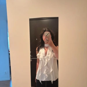 Vit volangblus från Zara - Supersnygg vit blus från Zara med volangdetaljer framtill och längs axlarna. Blusen är ärmlös, har hög hals med cut out och djup v-ringning. Luftigt material som känns lätt och fräscht, perfekt för en trendig look. Aldrig använd bara provad🫶🏻