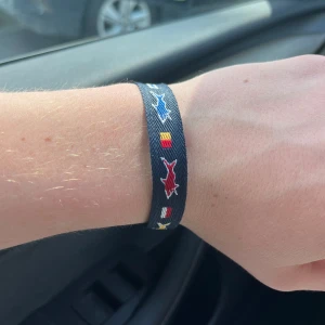 Marint tygarmband med flaggor - Snyggt marinblått tygarmband med broderade färgglada flaggor och fiskar i rött, blått, gult och vitt. Armbandet har justerbar knytning för perfekt passform och ger en cool nautisk vibe.