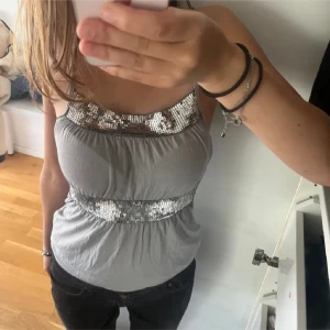 Grå paljettlinnetopp med smala axelband - Snygg grå linne-topp med smala axelband och breda silverpaljetter både vid bysten och midjan. Toppen har en lätt rynkad passform och är Superfin till svarta jeans eller kjol!