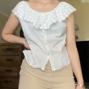 Vit blus med spetsvolang - Superfin vit blus med bred spetsvolang runt hela ringningen. Ärmlös modell med knappar framtill och lätt figursydd passform. Perfekt för en romantisk look och passar till både jeans och kjol. Luftigt bomullsmaterial som känns fräscht och somrigt. Skulle säga att det är en loose fit M.