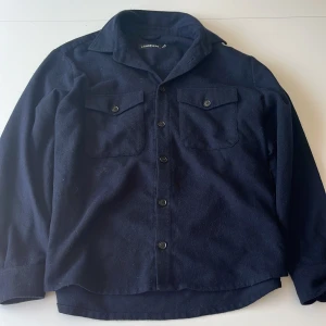 Mörkblå overshirt från J.Lindeberg - Stilren mörkblå overshirt från J.Lindeberg i ullmix. Skjortan har klassisk krage, två bröstfickor med lock och knappar framtill. Perfekt lager-på-lager-plagg med en clean look och normal passform.