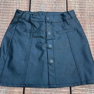 Svart skinnkjol med knappar framtill - Snygg svart kort skinnkjol med knappar längs hela framsidan. Kjolen har två dekorativa fickor framtill och är tillverkad i ett mjukt skinnliknande material. Perfekt för dig som vill ha en edgy och trendig look.