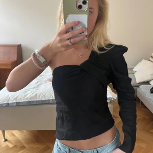 Svart oneshoulder topp med drapering - Säljer en svart oneshoulder topp med snygg drapering över axeln. Toppen har en lång ärm på ena sidan och är ärmlös på andra.