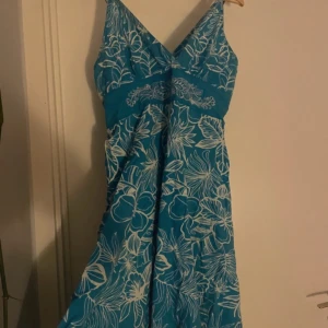 Blå blommig sommarklänning - Snygg blå klänning med vita blommor och bladmönster. V-ringad med breda axelband och markerad midja med broderad detalj. Lätt och luftig modell, perfekt för varma dagar. Klänningen har en avslappnad passform och är knälång.